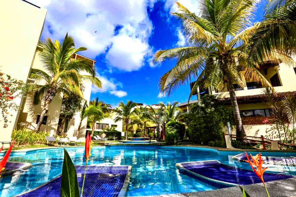 A shimmering pool is framed by lush palm trees, creating a tropical ambiance. The calm water reflects clear blue skies and white clouds, while comfortable loungers are scattered around the pool area, inviting relaxation near vibrant greenery and colorful tropical plants.