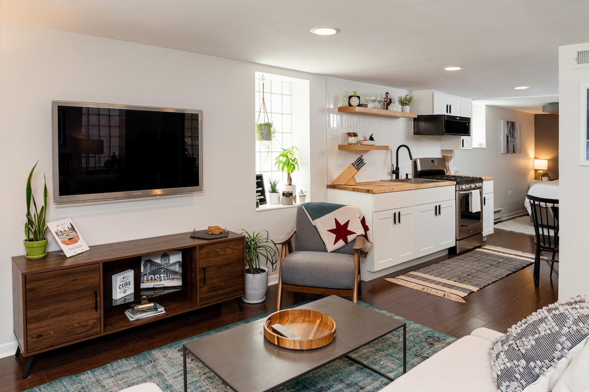 The living area features a sleek television mounted on the wall, complemented by a stylish wooden cabinet. A light-colored sofa is positioned in front of a rectangular coffee table, with a decorative bowl resting on it. The space is enhanced by natural light from the windows and greenery.