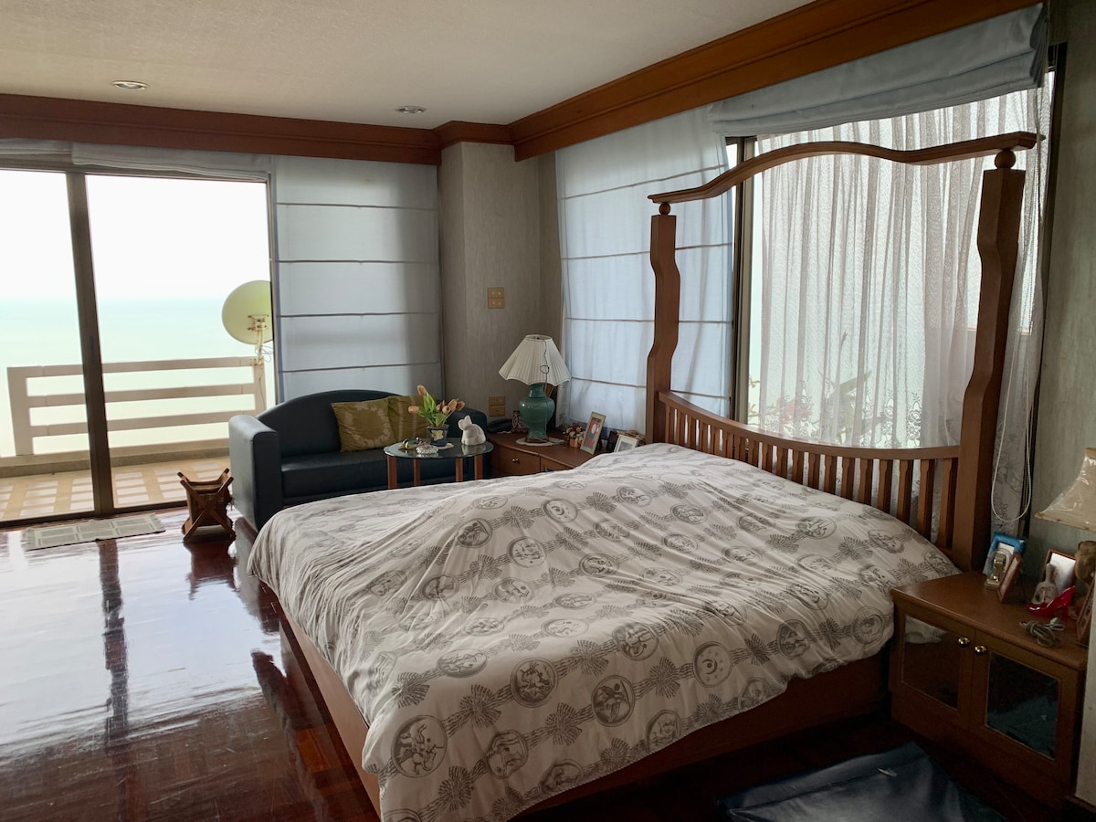 A spacious bedroom features a large wooden bed with patterned bedding. Natural light flows through sheer curtains, illuminating the room. A small seating area with a couch and decorative items is positioned near the balcony doors, which offer a view of the ocean.