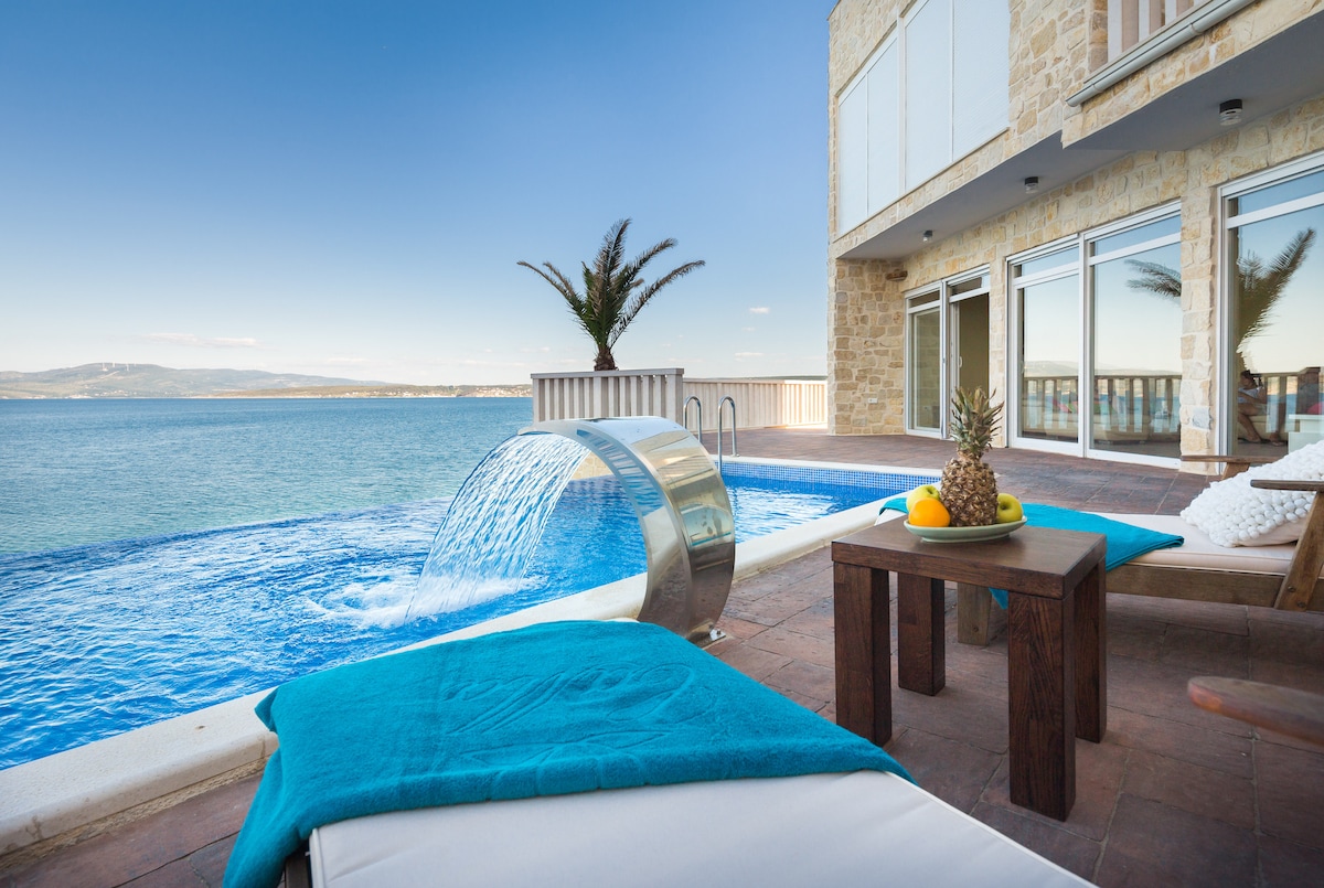 An inviting infinity pool is seen flowing into the sea, creating a seamless view. A lounge chair is positioned nearby, draped with a turquoise towel. A wooden table holds fresh fruit. The backdrop features a clear blue sky and tranquil waters.