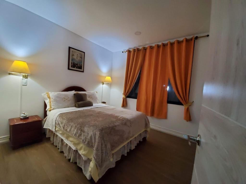 The bedroom features a spacious bed adorned with a light-colored bedspread, complemented by decorative pillows. Warm lighting is provided by bedside lamps and orange curtains frame the window, enhancing the cozy ambiance.