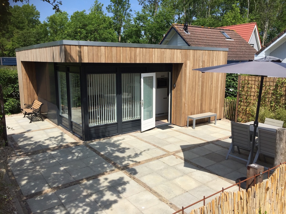 A modern, single-story house is featured, showcasing natural wooden siding and large glass windows. A patio area with seating and an umbrella provides outdoor relaxation. The entrance includes a white door, flanked by vertical blinds that allow natural light to fill the interior.