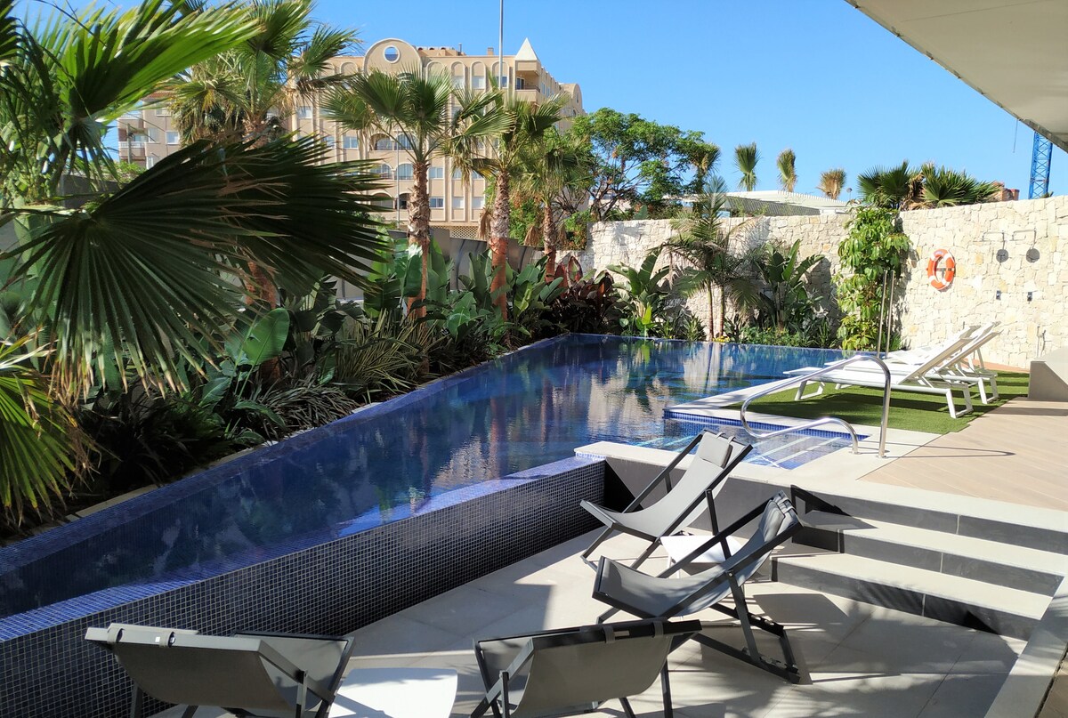 A tranquil pool area is surrounded by lush tropical plants, featuring a shimmering blue pool and several lounge chairs. The sleek pool design complements the natural surroundings, and warm sunlight enhances the inviting ambiance of the outdoor space.