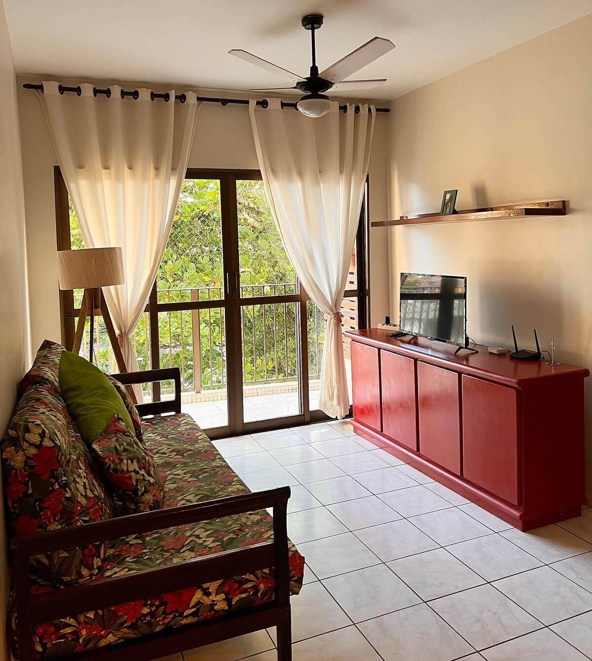 A bright living area is presented with large windows covered by sheer curtains, allowing natural light to fill the space. A colorful sofa with floral patterns is paired with a wooden coffee table. A television is mounted on a red shelf, providing a casual entertainment area.