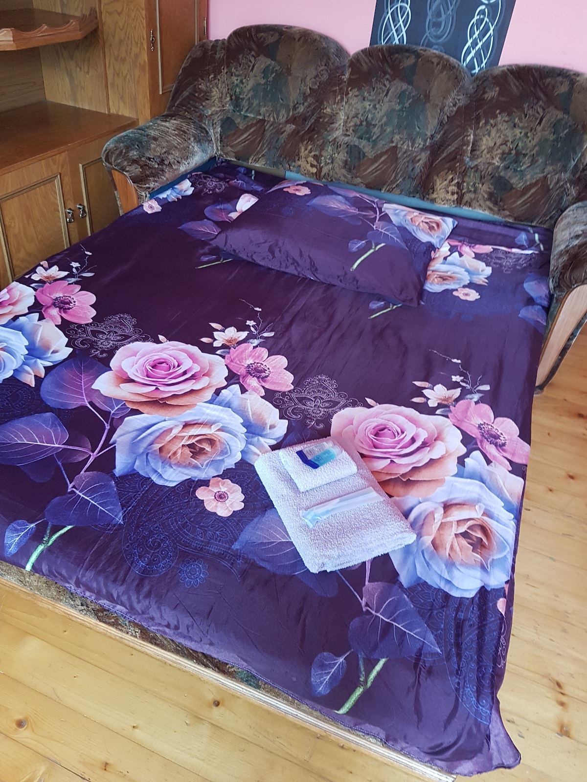 A floral-patterned blanket covers a spacious sofa bed, with additional decorative pillows arranged on top. Nearby, a towel is neatly placed, complementing the vibrant colors of the bedding. The surrounding wooden cabinetry and wall add warmth to the sleeping area.
