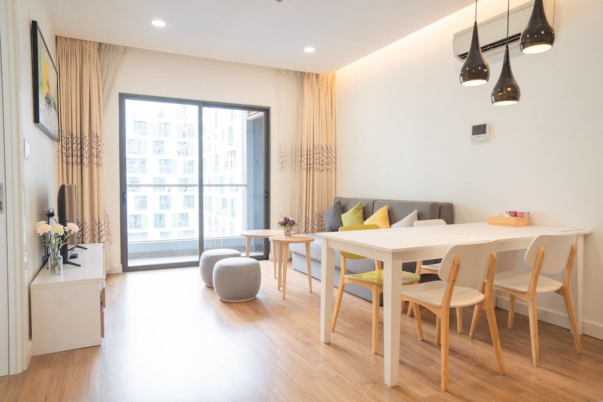 A modern living area features a light-colored sofa adorned with decorative cushions. A dining table with wooden chairs and a small ottoman are present. Natural light shines through the large glass doors leading to a balcony, enhancing the spacious feel of the room.
