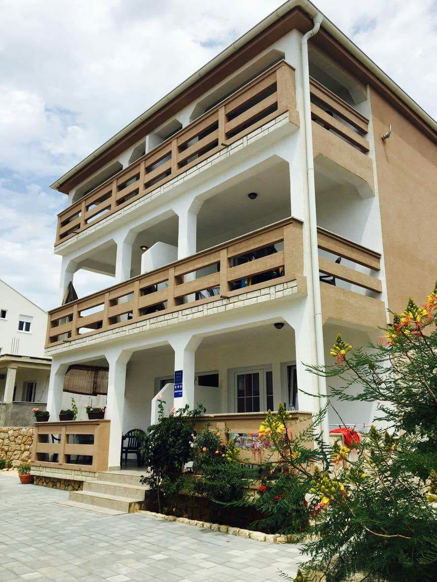 A three-story building is displayed, featuring large balconies with railings on each level. The exterior is characterized by a neutral color palette and minimalistic design. A landscaped area with greenery is visible in the foreground, accompanied by stone pathways leading to the entrance.