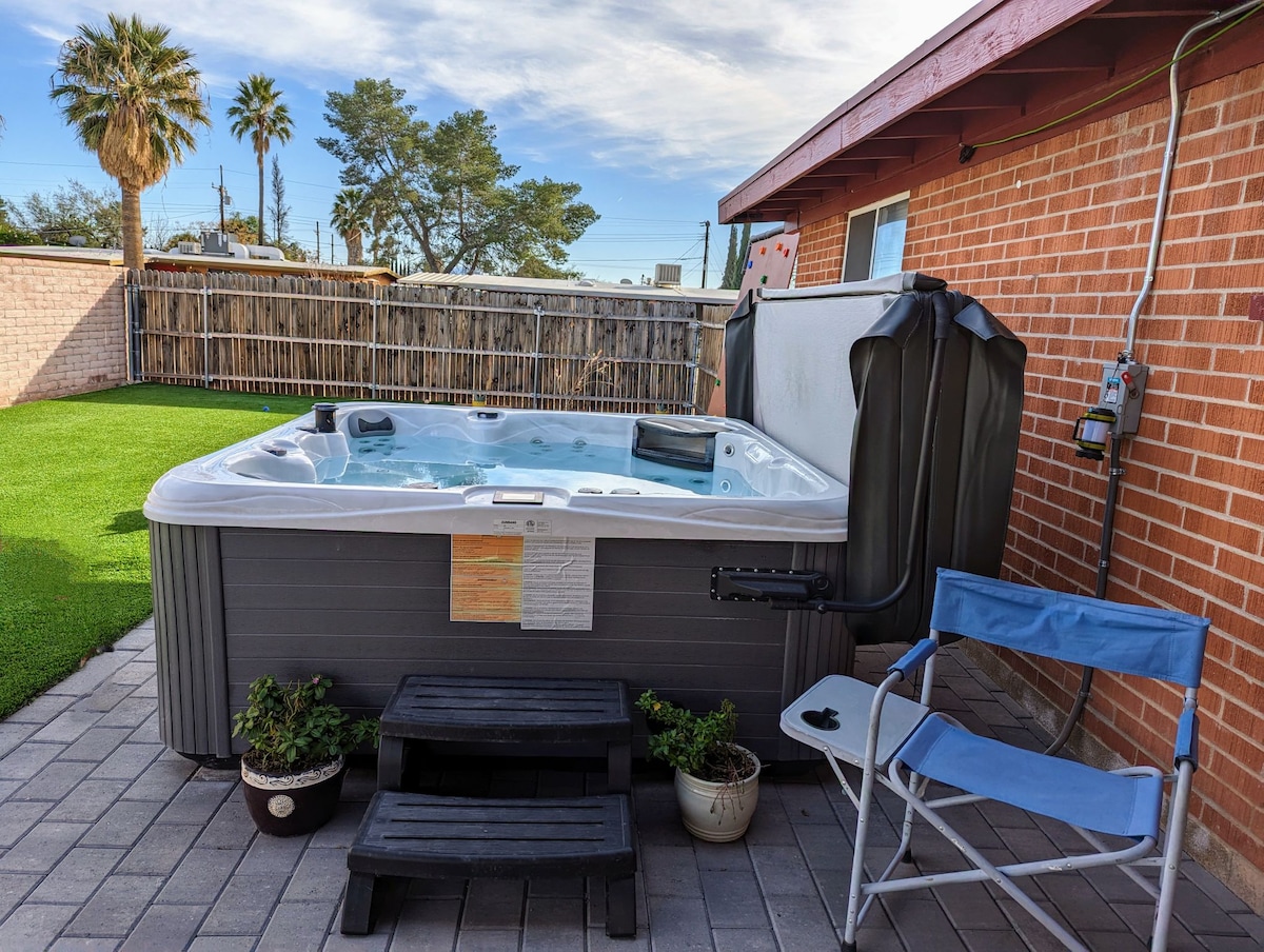 A hot tub with built-in seating is surrounded by potted plants on a paved patio. Grass outlines the area, creating a green space. A chair is positioned nearby, and a brick wall adds privacy, with palm trees visible in the background.