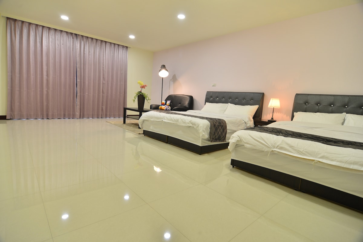 A light-filled room features two spacious beds with crisp white linens and dark headboards. A sleek black sofa and a bedside lamp add to the comfort. Floor-to-ceiling curtains frame the window, while polished tiles create a clean and airy atmosphere.