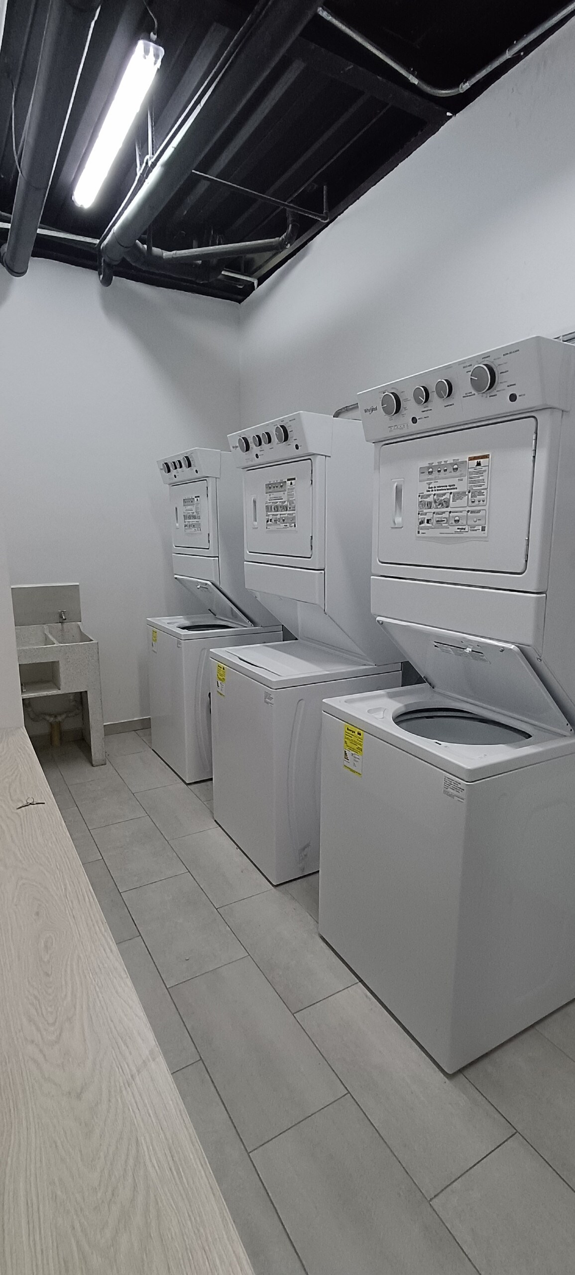 The communal laundry area features three stacked washer-dryer units against a light-colored wall. A small sink is positioned beside the appliances. Neutral tile flooring extends throughout the space, providing a clean and functional environment for laundry tasks.