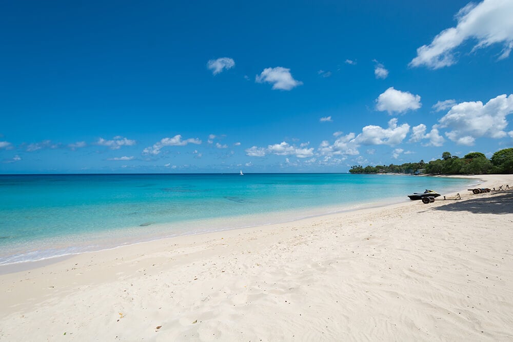 A tranquil beach scene is displayed, featuring soft, white sand meeting calm turquoise waters. Gentle waves lapping against the shore are highlighted under a clear blue sky, dotted with a few fluffy clouds. Sun-loungers are visible along the beach, inviting relaxation.