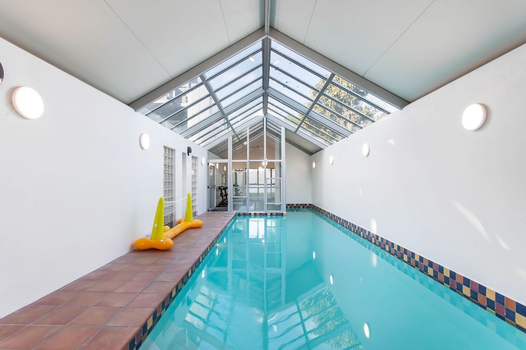 An indoor swimming pool is presented with a clear, reflective surface under a transparent roof, allowing natural light to fill the space. Two colorful inflatable loungers are visible beside the water, enhancing the bright and relaxing atmosphere of the pool area.