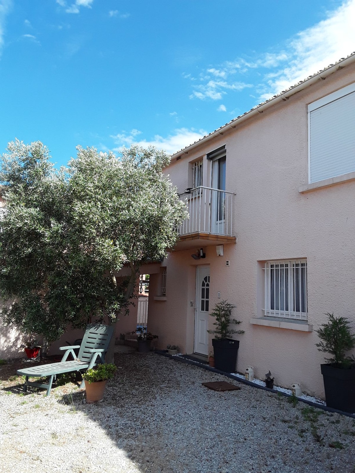 The exterior of a two-story residence is depicted, featuring peach-coloured walls complemented by a light blue sky. A small balcony is visible above the entrance door, alongside a neatly landscaped area with potted plants. A couple of lounge chairs are positioned on a gravel pathway.