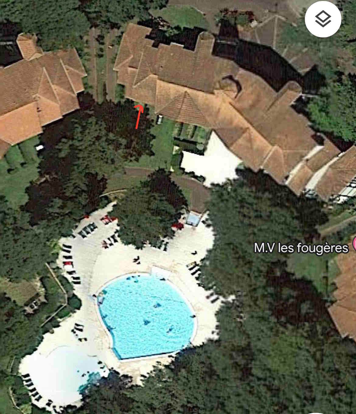 An aerial view showcases a well-maintained residential area featuring several buildings surrounded by lush greenery. A large swimming pool is centrally located, with loungers arranged around its perimeter. Pathways connect the individual residences to the pool area.