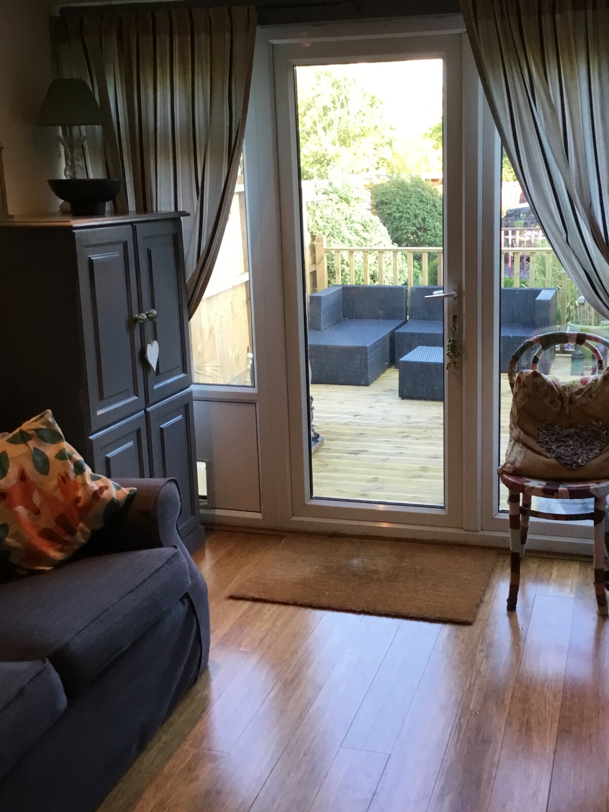 A seating area features a gray couch adorned with a colorful decorative pillow. Large glass doors open to a sunlit wooden deck, which includes modern outdoor seating. Natural light enhances the inviting ambiance, while partially drawn curtains frame the view.