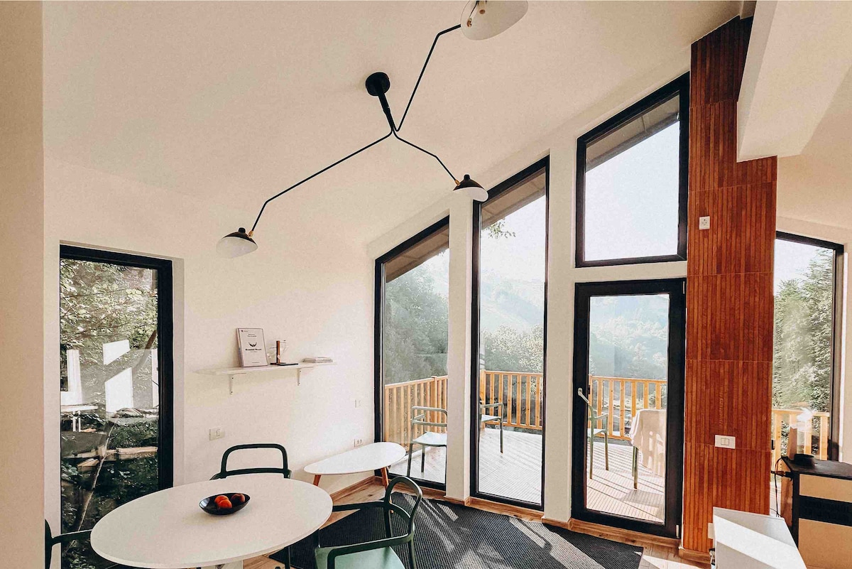 A bright open-space area features large windows that invite natural light and offer views of the surrounding trees. A round table and chairs are arranged next to the glass doors, which lead to an outdoor deck. Modern lighting hangs from the ceiling.