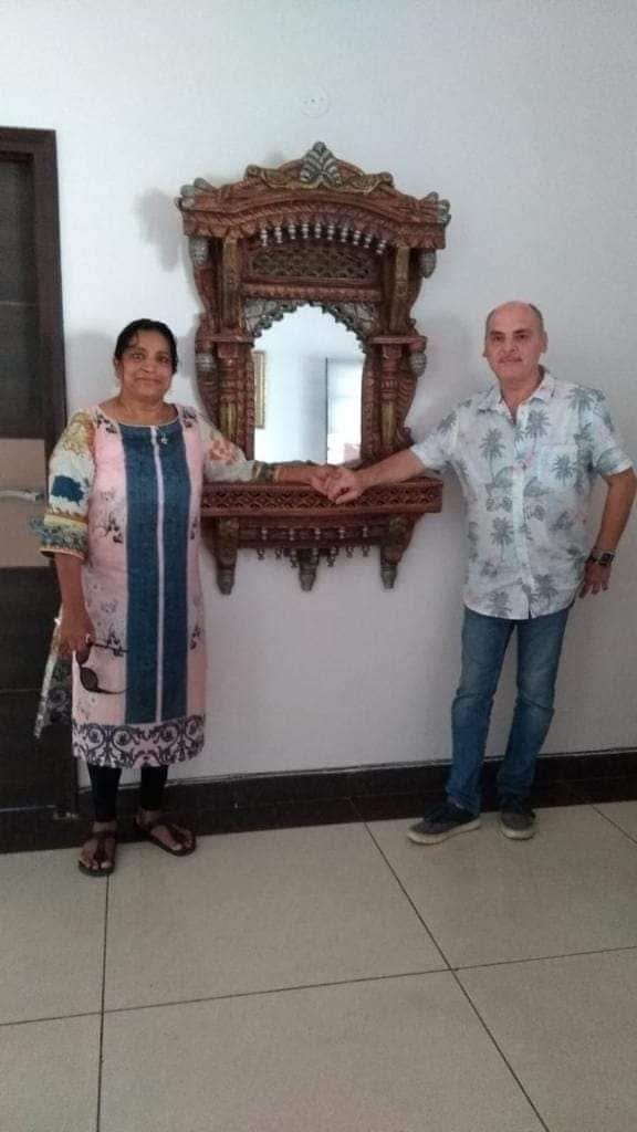 A decorative mirror with intricate carving is mounted on a light-colored wall. Two individuals are positioned in front of the mirror, each engaged in a handshake. The floor features large square tiles, reflecting a neutral color palette.