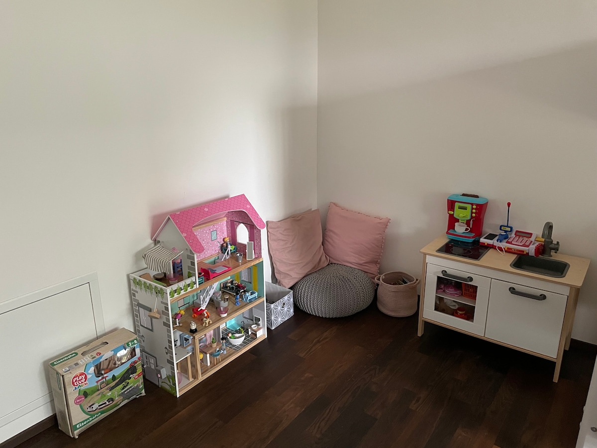 A play area is seen with a well-organized toy shelf featuring a dollhouse and various playsets. Cozy cushions are arranged in the corner, complemented by a small play kitchen set. Light wood flooring enhances the inviting atmosphere.