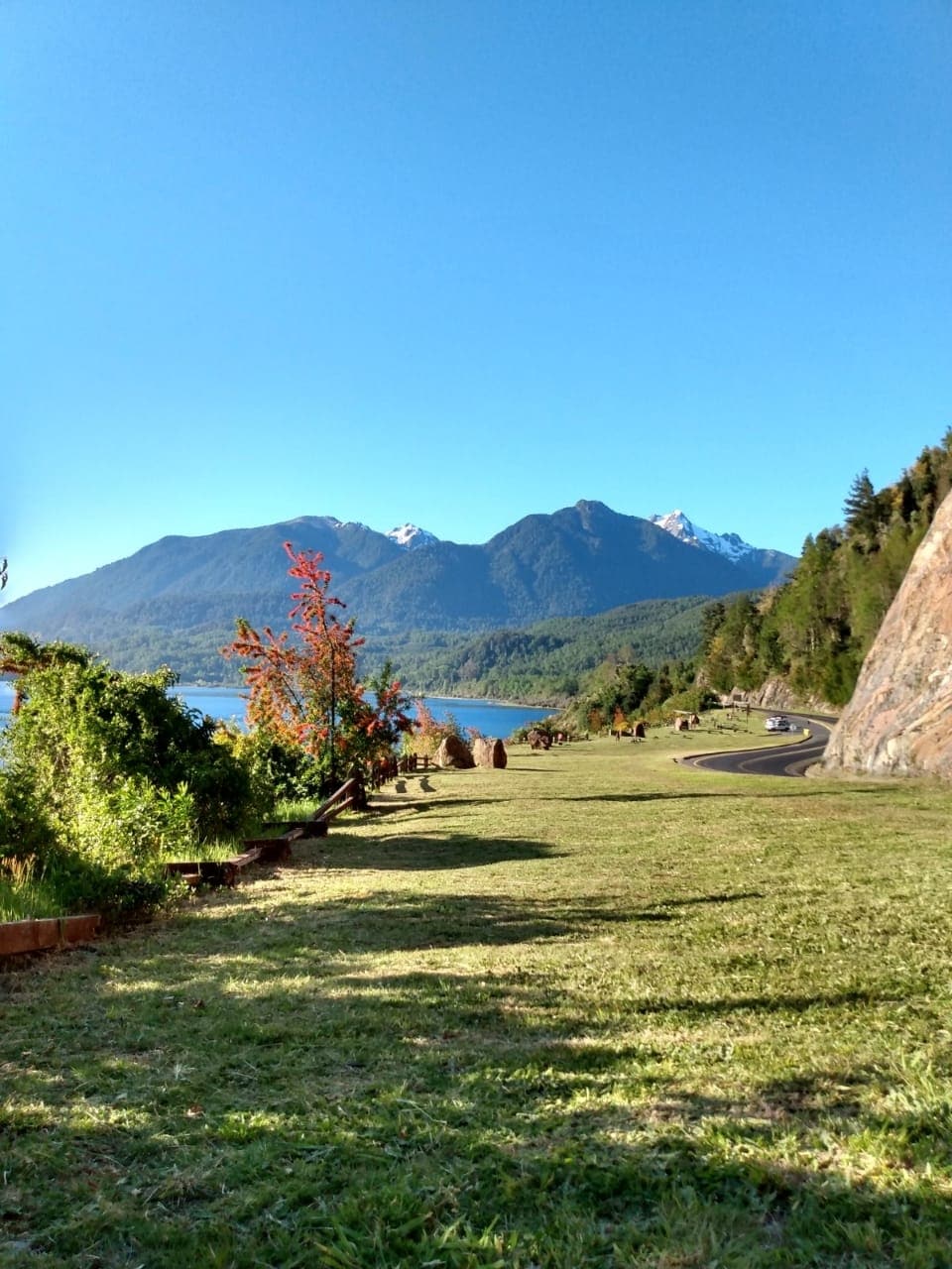 A scenic outdoor area is displayed, featuring a grassy expanse bordered by trees. Mountains are visible in the distance, partially covered by snow, while a clear blue sky completes the serene landscape. A winding road can be seen alongside the area.