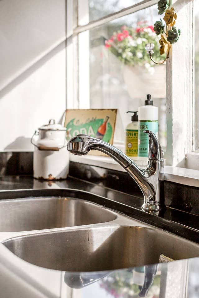 A stainless steel sink is positioned in front of a window, showcasing bright greenery outside. The counter includes a bottle of dish soap and a decorative vintage sign. Natural light illuminates the scene, enhancing the functional kitchen space.