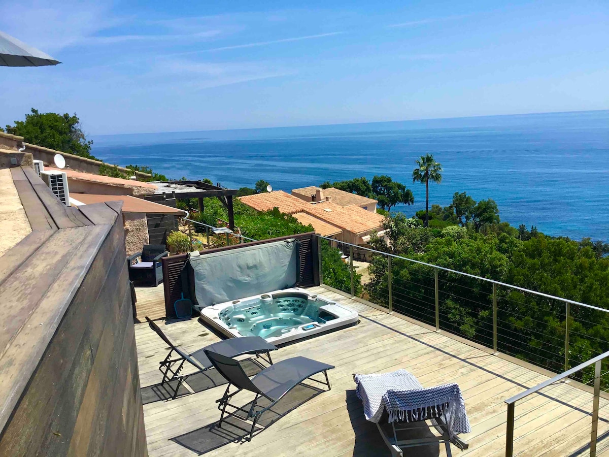 A spacious terrace offers a jacuzzi with panoramic ocean views. Sun loungers are positioned nearby, inviting relaxation under the clear sky. The natural surroundings feature lush greenery and distant rooftops, highlighting a serene coastal atmosphere.