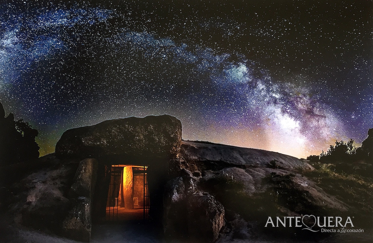 A night scene depicts an ancient dolmen illuminated by soft light. The structure, made of large stones, is framed by a starry sky featuring the Milky Way. The contrast between the warm glow from inside the dolmen and the cool tones of the surrounding darkness is striking.