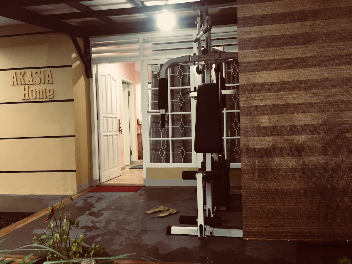 A fitness area is visible, featuring a multi-gym equipment set against a textured wall. The entrance to the home is framed by a white door adorned with glass panels, leading to a warmly lit interior space. Potted plants add a touch of greenery.