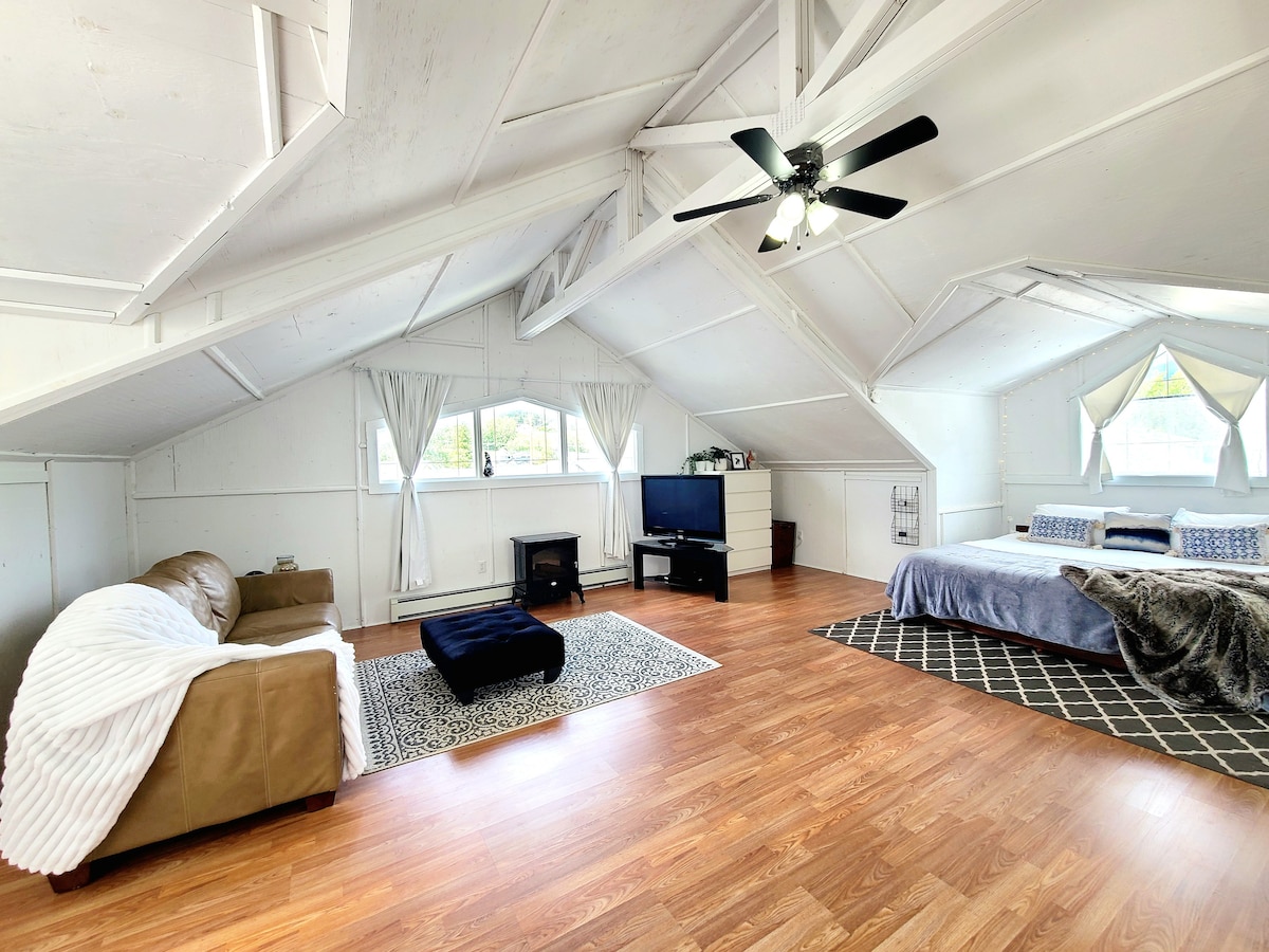 A spacious loft area features an inviting lounge space, including a sofa and a dark ottoman. A large bed is complemented by light-colored bedding. Natural light filters through several windows, creating a bright atmosphere. A television is positioned opposite the seating area.
