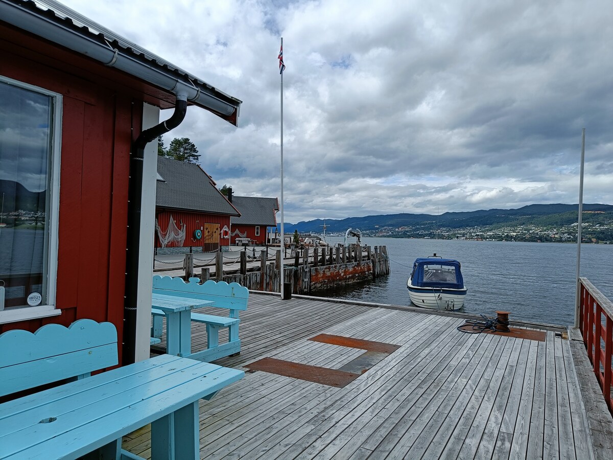 A wooden deck extends towards the water, featuring a light blue picnic table. A small boat is visible moored at the dock, with a gently waving Norwegian flag nearby. The background showcases a scenic view of the shoreline and mountains under a partly cloudy sky.