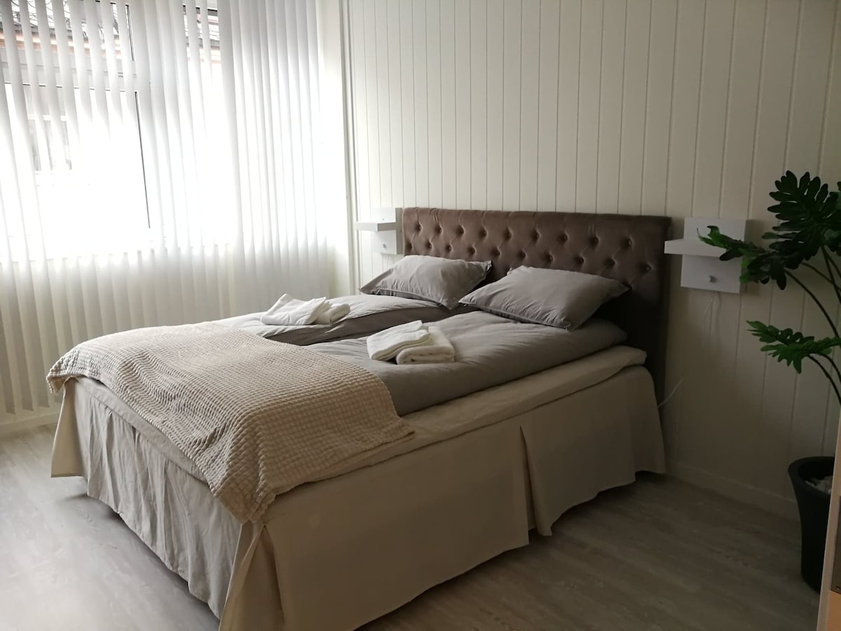 A spacious bed is presented with soft linens and two neatly placed towels. Natural light enters through vertical blinds, illuminating the room. Adjacent to the bed, minimalistic nightstands provide convenient surfaces, while a potted plant adds a touch of greenery.