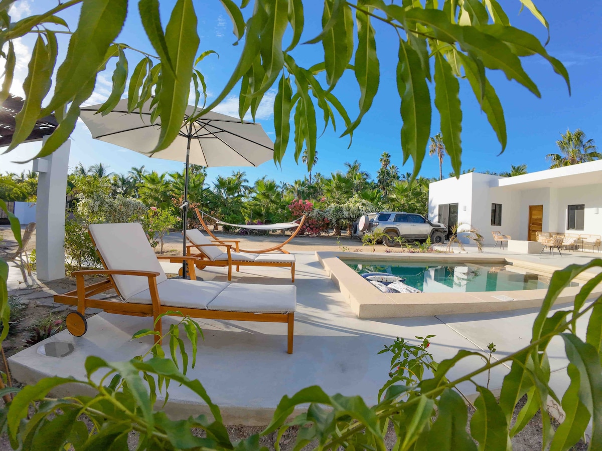 A relaxing outdoor space features a pool framed by a clear blue sky and palm trees. Sun loungers and a large umbrella offer shaded seating. A hammock is visible nearby, providing an inviting spot to unwind amidst the lush greenery surrounding the area.