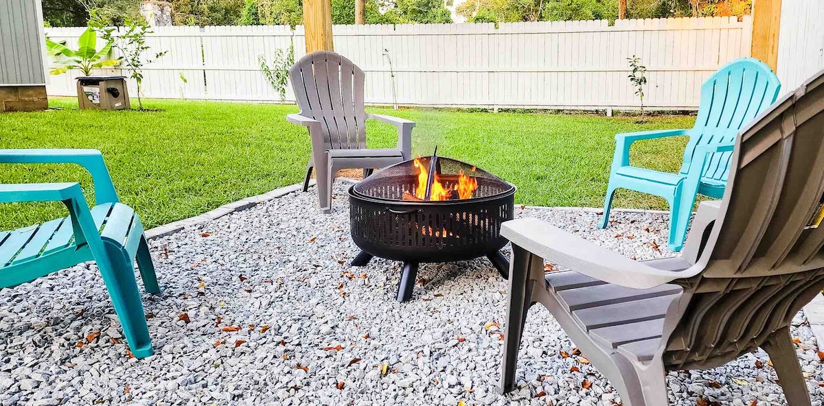 A firepit is centered in a gravel area, surrounded by four brightly colored chairs. The fenced backyard features a neatly manicured lawn, providing a comfortable outdoor setting for relaxation and social gatherings.