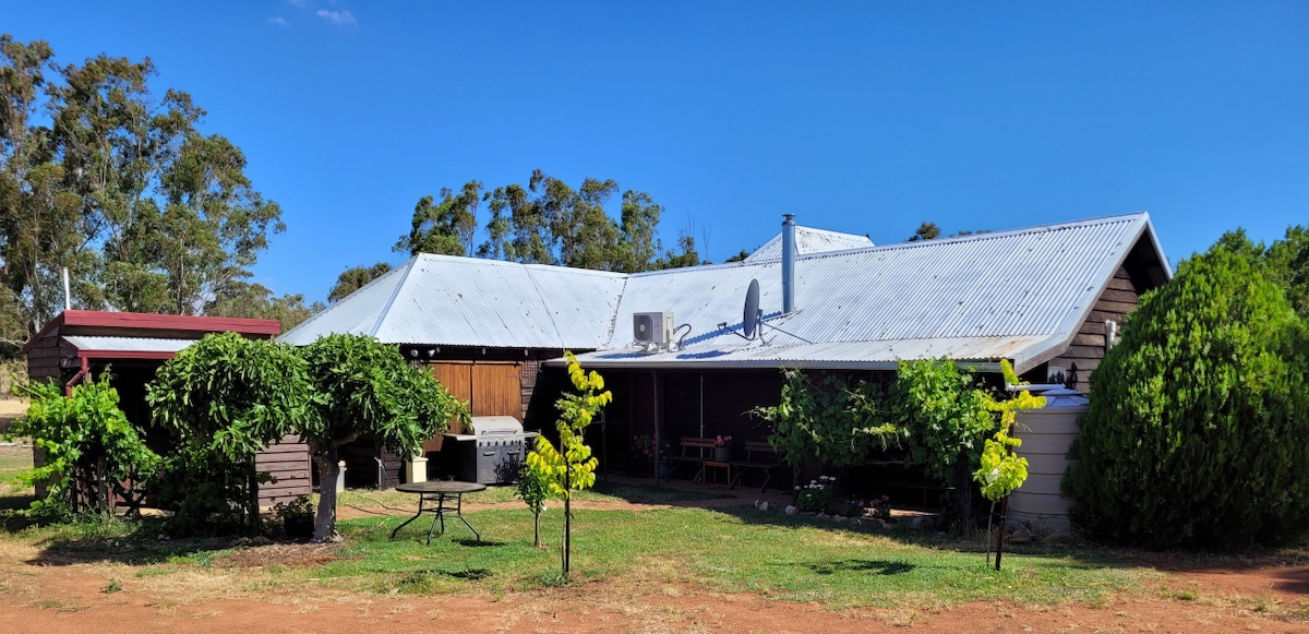 The rustic coachhouse features a metal roof and wooden siding, surrounded by green grass and young trees. Outdoor seating and a table are visible, providing options for relaxation. Clear blue skies and gentle hills in the background enhance the rural setting.