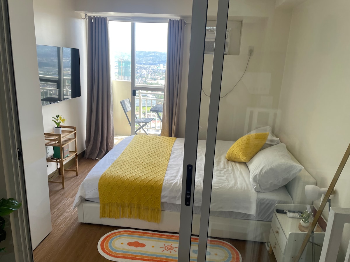 A cozy bedroom features a spacious bed adorned with white linens and a sunny yellow throw. A large window provides a view of the city, framed by light curtains. A small table and chairs are positioned near the window, next to a decorative plant.