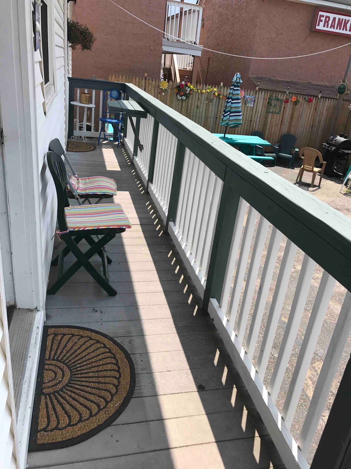 A narrow balcony area is highlighted by two chairs with striped cushions, set against white railings. The floorboards are sunlit, and a welcome mat is visible near the entrance. In the background, colorful furniture can be seen, creating a casual outdoor environment.