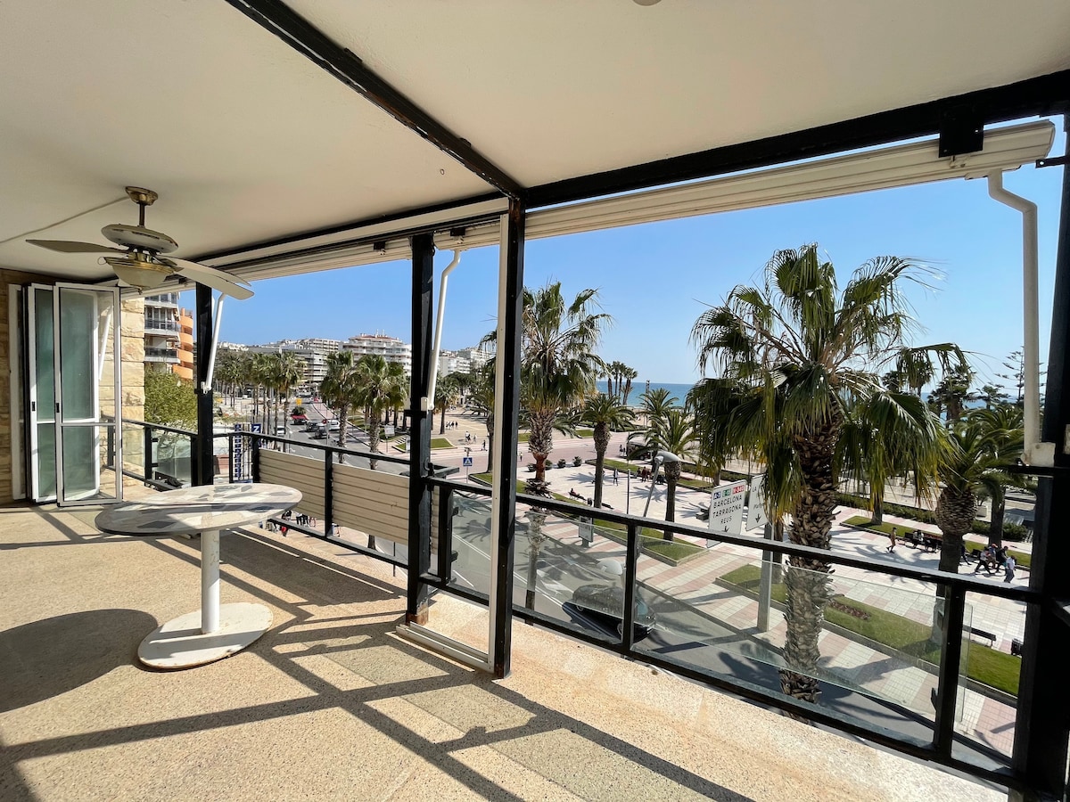 A spacious balcony offers a clear view of palm trees and ocean front. Sunlight fills the area, which includes a round table and ceiling fan. Floor-to-ceiling glass doors provide an unobstructed connection to the vibrant outdoor scenery.