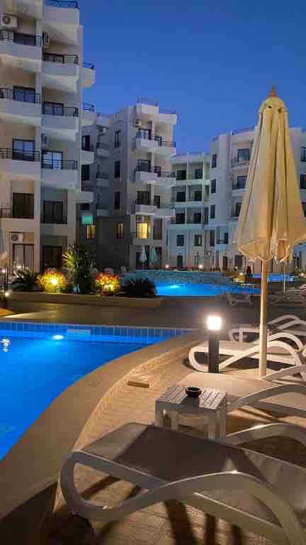 Illuminated swimming pools are surrounded by modern white buildings, creating a serene atmosphere. Sun loungers positioned near the water’s edge offer relaxation options. A large parasol provides shade, while soft lighting enhances the evening ambiance.