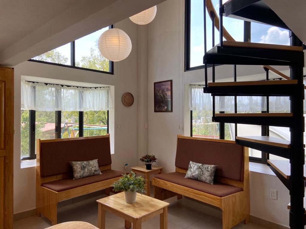 A cozy seating area is presented with wooden benches and a light wooden table. Natural light fills the space through large windows, and paper lanterns hang from the ceiling. A spiral staircase is visible, leading to the upper level.