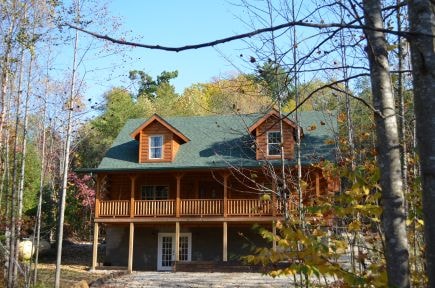 A log cabin stands gracefully among trees, featuring a green roof and a spacious front porch with wooden railings. Large windows reflect natural light, while surrounding foliage displays a mix of green and autumn colors.