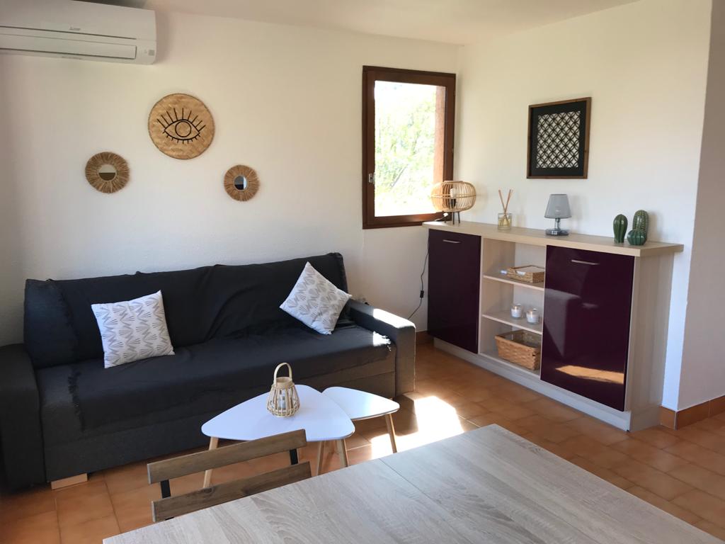 The living area is furnished with a dark sofa featuring decorative pillows. A small coffee table sits in front, with a sideboard displaying decorative items. Natural light enters through a window, and wall art complements the space. An air conditioning unit is mounted above.