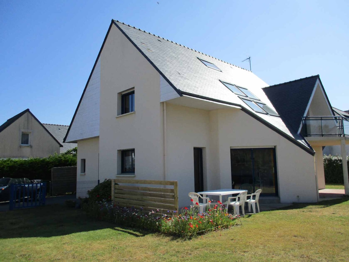 A modern villa with a sloped roof is displayed against a clear blue sky. Large windows enhance natural light in the living spaces. A patio area includes a table and chairs, while a small garden features colorful flowers, contributing to the tranquil outdoor setting.