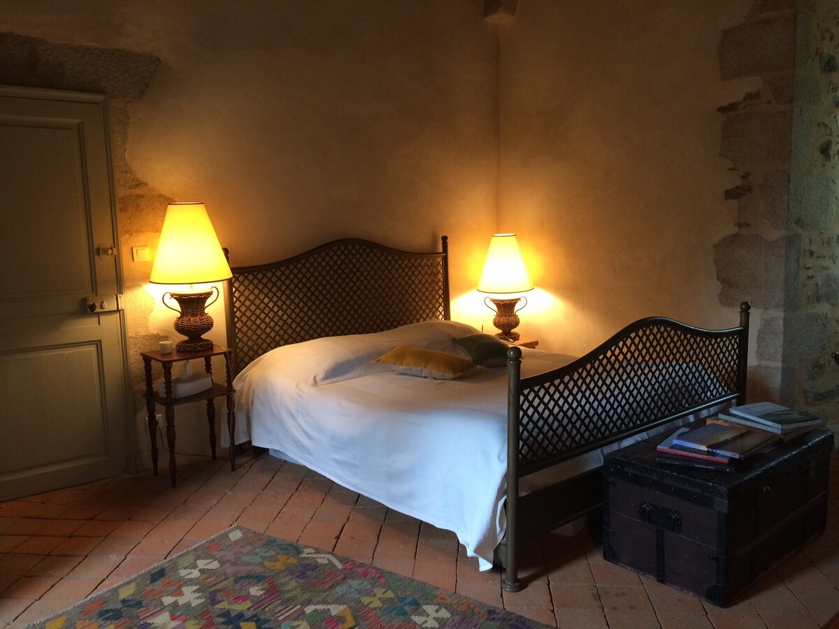 The room features a dual bed with a woven metal headboard, flanked by two bedside lamps emitting a soft glow. A colorful rug lays over terracotta tiles, while a small chest and a stack of books are positioned at the foot of the bed. Textured stone walls add character.