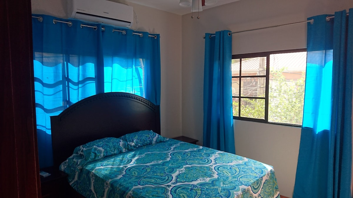 A well-lit bedroom features a spacious bed adorned with a colorful patterned bedspread. Large blue curtains frame a window that allows natural light to fill the room, while light-colored walls create a bright and airy feel.
