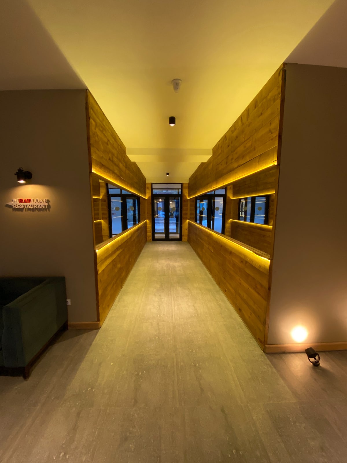 A long corridor is illuminated with soft lighting along wooden walls. Large windows on either side provide views of the outside. The floor is finished in a smooth surface, leading to a glass entrance door at the end of the hallway.