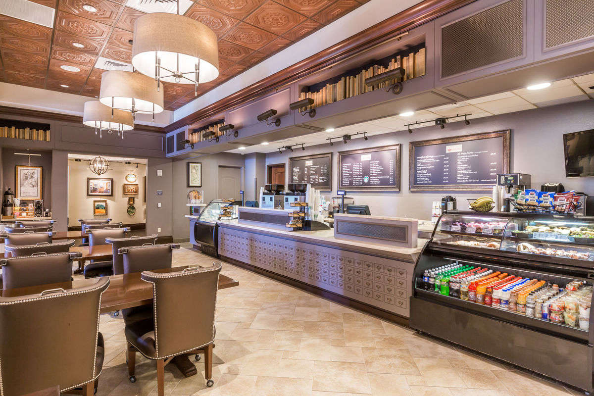 A well-appointed café space features stylish brown chairs and tables arranged for comfort. The counter displays a variety of snacks and beverages. Soft lighting complements the detailed ceiling and wall art, creating a welcoming environment for guests to relax and enjoy.