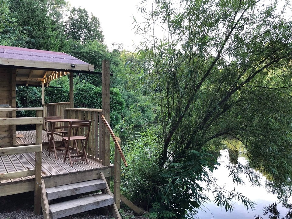A wooden deck area is positioned beside a tranquil lake, featuring a small table and two chairs. Surrounding greenery includes tall trees and shrubs, creating a peaceful natural setting. Steps lead down from the deck towards the water, enhancing accessibility.