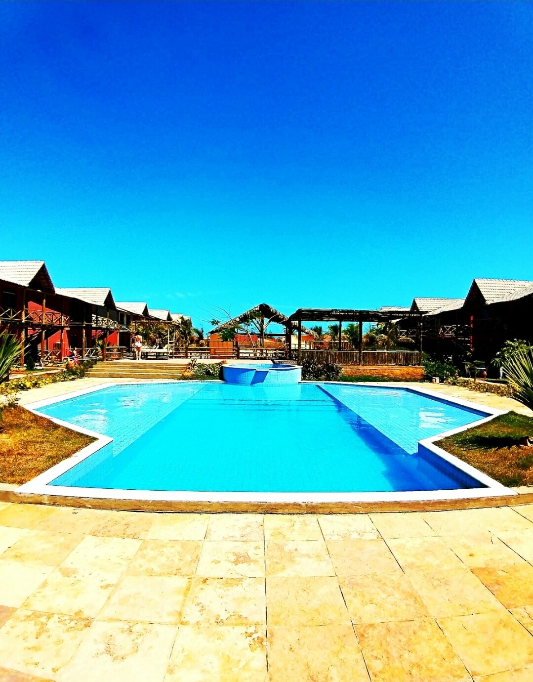 A clear blue swimming pool is centrally located in the outdoor area, surrounded by landscaped gardens. Comfortable lounge spaces and palm trees are visible in the background, offering an inviting setting. The sky is bright and clear, enhancing the atmosphere of relaxation.