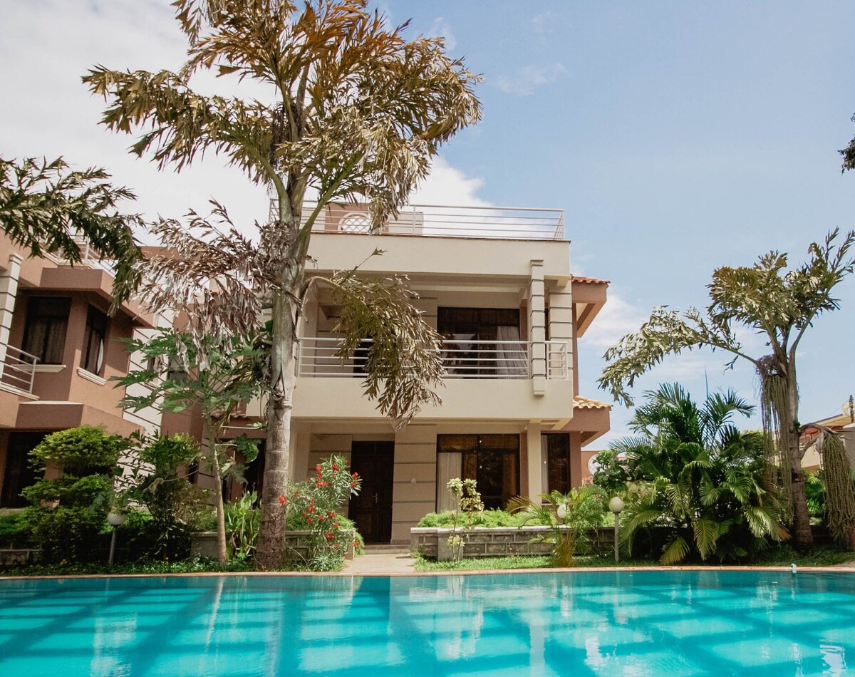 The villa stands gracefully amidst lush greenery, with a well-maintained garden visible in the foreground. The spacious two-story structure features large windows and balconies overlooking the shimmering pool, reflecting the serene surroundings and clear blue sky.