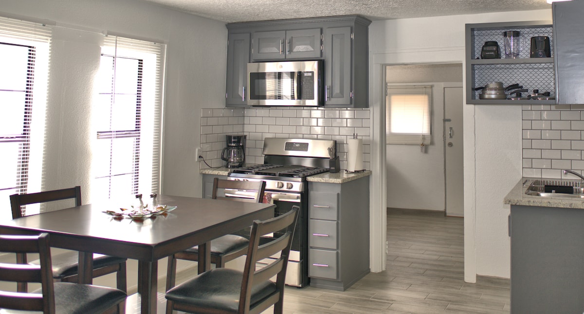 The kitchen area features modern cabinetry and appliances, with a dining table set for four. Natural light enters through multiple windows, illuminating the sleek countertops. A doorway leads to another part of the home, enhancing the open layout.