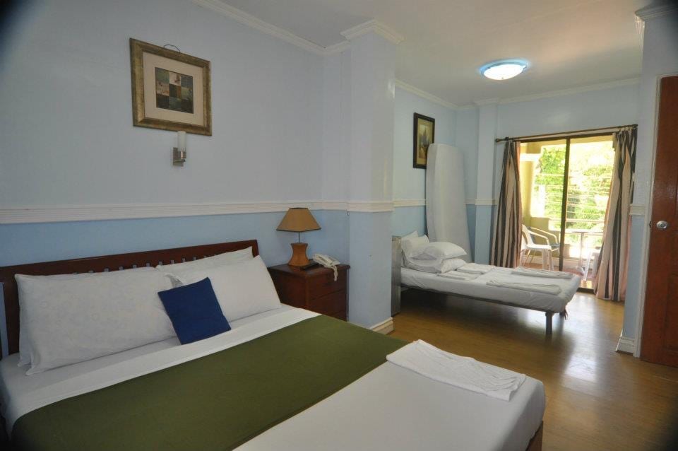 A spacious room features a large bed with a decorative pillow and a smaller bed beside it. A wooden nightstand holds a lamp. Light enters through the glass door leading to a balcony, which provides a view of outdoor greenery.